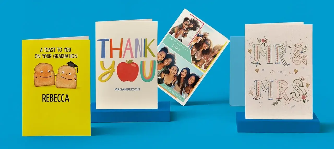 'The image shows four greeting cards displayed on blue stands against a blue background. From left to right: a yellow graduation card with smiling toast characters and the text 'A TOAST TO YOU ON YOUR GRADUATION REBECCA'; a thank you card with colorful 'THANK YOU' letters and an apple illustration labeled 'MR SANDERSON'; a photo collage titled 'FAMILY FUN'; and a white wedding card with decorative 'MR & MRS' text surrounded by hearts and floral designs.'