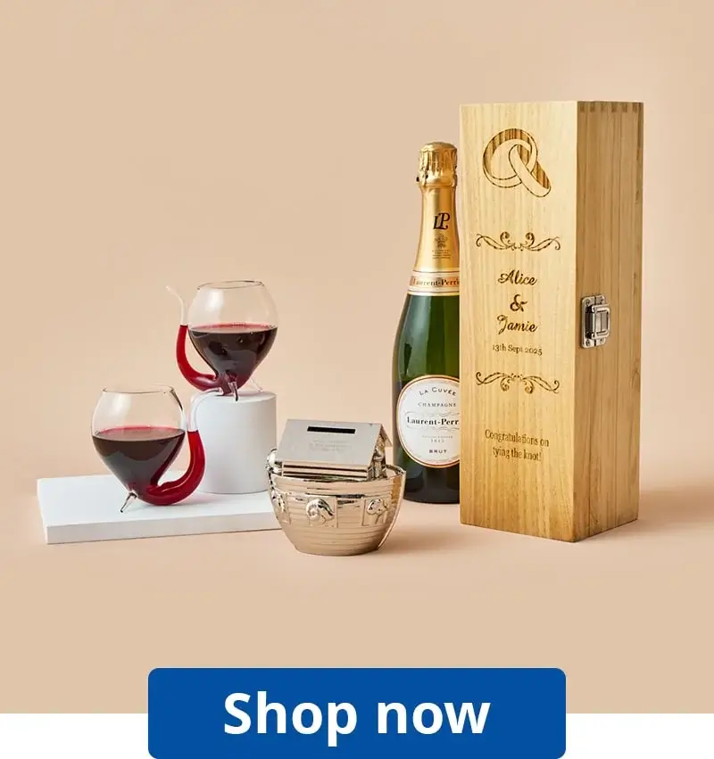 The image shows a gift set that includes a bottle of champagne, two wine glasses with red wine, a wooden box with engraved text, and a small silver container. The wooden box has an engraving of two interlocking rings at the top, followed by the names 